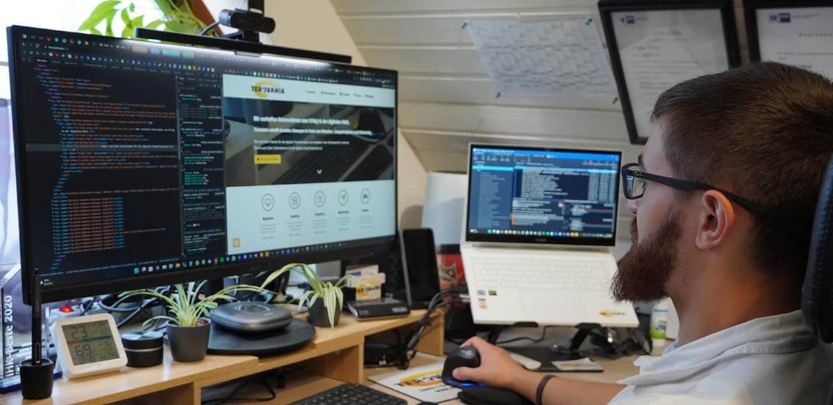 A web developer in a casual white shirt working across multiple monitors displaying code and the Terovania website. His workspace is organized with coding environments on two large screens, showcasing active development and website management. The setup includes various office tools and plants, reflecting a professional and efficient work environment.