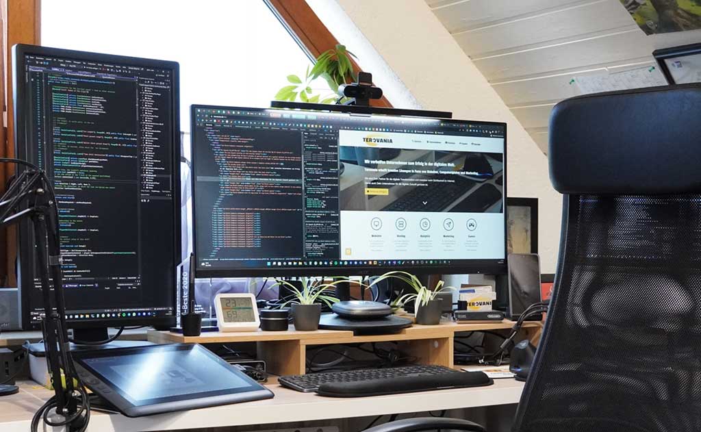 A web developer's workspace with a vertical monitor displaying code, and a third monitor showing the Terovania Solutions website homepage. The desk is equipped with various tech accessories like a drawing tablet, headphones, and indoor plants.